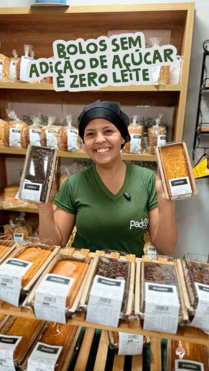 Atendente da Padoca sorrindo ao lado da prateleira com a placa Bolos sem adição de açúcar e zero leite