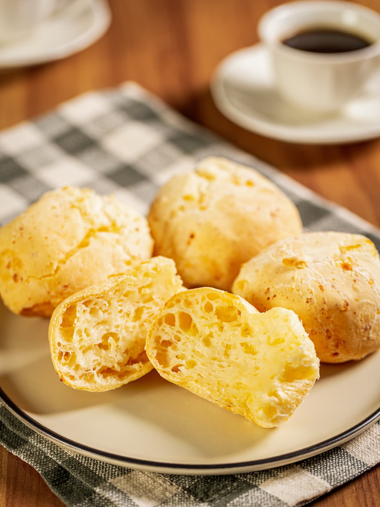 Pão de queijo dourado num prato com uma xícara de café sobre toalha xadrez verde e branca