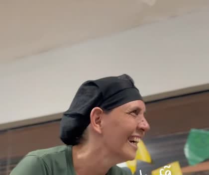 Rosa, atendente da Padoca, rindo de camisa verde e touca preta atrás do balcão